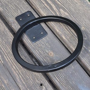 May include: A matte black metal ring holder featuring a flat rectangular mounting plate with four pre-drilled screw holes. The circular ring is welded to the center of the plate, creating a sturdy, functional hardware piece for wall mounting.