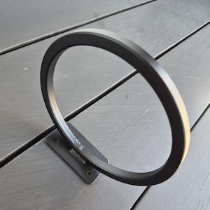 May include: A black metal ring attached to a rectangular base with two screw holes. The ring is circular and stands upright, creating a minimalist design. The object is set against a dark gray wooden surface.