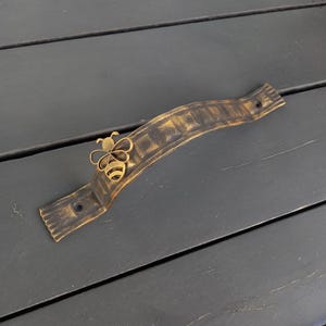 Op de afbeelding: Een decoratieve, gebogen metalen handgreep met een bijenaccent. De handgreep heeft een donker bronzen kleur met een textuur. De bij is goudkleurig en is in het midden van de handgreep bevestigd. De handgreep heeft twee montagegaten.