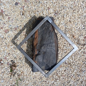 May include: A silver metal square frame sits on a bed of small white stones. The frame is empty, but a dark gray rock with a reddish brown streak is visible through the frame.