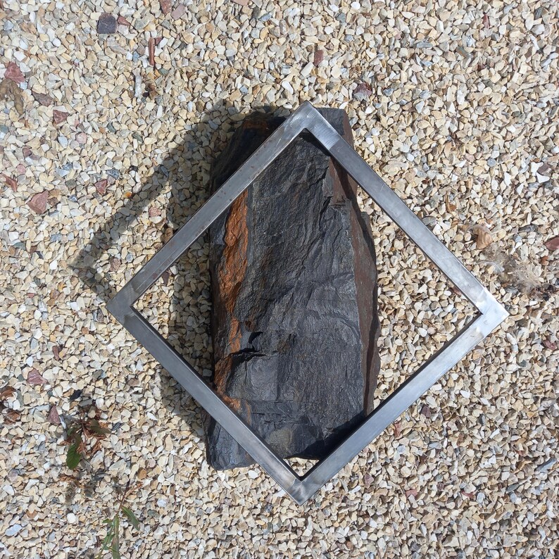 May include: A silver metal square frame sits on a bed of small white stones. The frame is empty, but a dark gray rock with a reddish brown streak is visible through the frame.