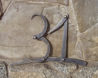 May include: A metal house number plaque with the number 31 in a stylized font. The plaque is attached to a stone wall.