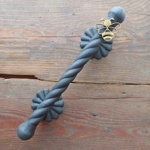 以下が含まれることがあります： ねじれたデザインと装飾的な蜂のアクセントが付いた黒い金属製のドアハンドル。ハンドルは木製の表面に取り付けられています。