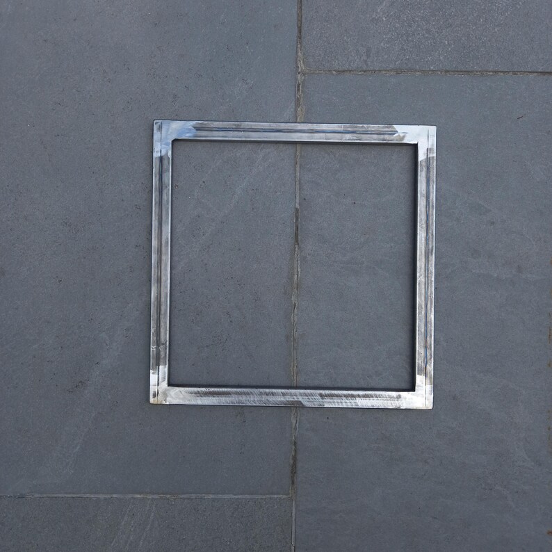May include: A silver metal square frame with a smooth, polished finish. The frame is set against a gray tiled background.