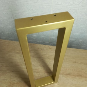 May include: A gold-coloured, rectangular table leg with a trapezoidal design. The leg has a hollow, open centre and three pre-drilled holes on the top surface for easy attachment. The leg is made of metal and has a smooth, matte finish.