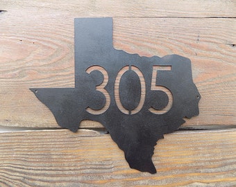 May include: Black metal Texas state outline with the number 305 cut out in the center. The outline is mounted on a wood background.