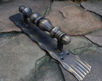 May include: A bronze-colored, forged metal door pull with a decorative handle. The handle has a series of rounded sections and is mounted on a rectangular backplate with a wavy edge. Two screw holes are visible.