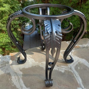 Può includere: Base di tavolo in metallo nero con decorazioni a volute e motivi a foglia. La base ha un piano rotondo e tre gambe.