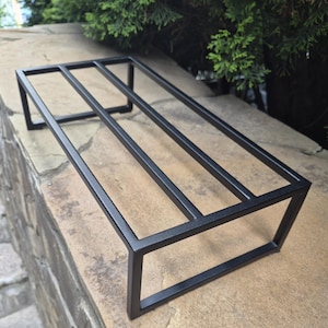May include: A rectangular black metal stand featuring a minimalist frame construction with four parallel horizontal bars supported by two vertical end pieces, creating a raised platform design with a clean, open, and geometric structure.