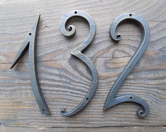 May include: A set of three metal house numbers, 1, 3, and 2, in a decorative, stylized font. The numbers are a dark bronze color and are mounted on a wooden surface.