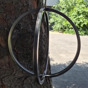 Può includere: Un ornamento sferico costruito con due anelli di metallo grigio scuro che si intersecano. La struttura è sospesa da un sottile cordino, creando una forma a globo geometrico che presenta una finitura opaca liscia sulle fasce metalliche.