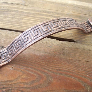 Op de afbeelding: Een koperkleurige metalen deurgreep met een decoratief Grieks sleutelpatroon.