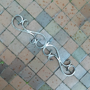 Può includere: Un pezzo decorativo in metallo argentato con un design a rotolo. Il pezzo è in metallo e ha una finitura lucida. È appoggiato su una superficie piastrellata.