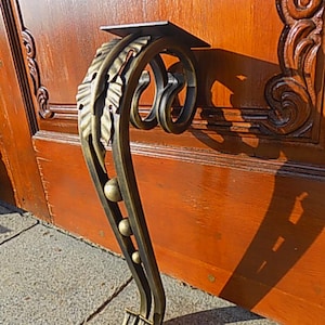 May include: A decorative metal furniture leg with a scroll design and a leaf motif. The leg is finished in a gold tone and has a black metal plate at the top.