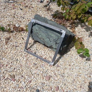 Könnte beinhalten: Ein Metallrahmen mit geschweißten Ecken, der auf einem Bett aus kleinen, hellen Steinen liegt.