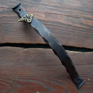 May include: A black, hand-forged metal handle with a butterfly accent. The handle has a textured, hammered appearance and is mounted on a wooden surface. The butterfly is a gold colour.