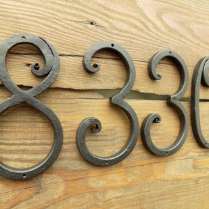 May include: Black metal house numbers with a decorative, scrolled design. The numbers are 8, 3, and 0.