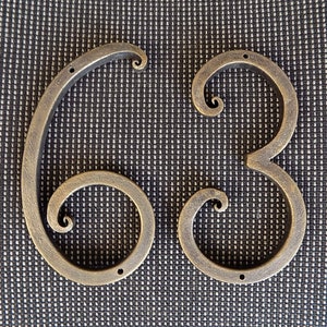 May include: Two metal house numbers, 6 and 3, are displayed against a black textured background. The numbers are made of dark bronze metal and have a decorative, swirling design.
