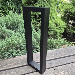 May include: A matte black metal furniture leg featuring a rectangular frame design. The structure is composed of four straight, solid bars joined at right angles to form a tall, open-centered vertical rectangle, providing a stable base for furniture.