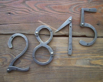 May include: Close-up of a set of metal house numbers, 2815, attached to a wooden surface. The numbers are made of dark metal and have a rustic, hand-forged look.