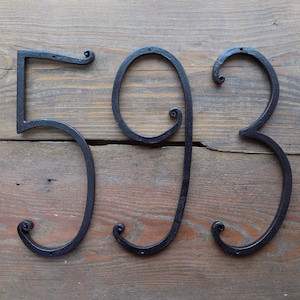 May include: Black metal house numbers 593 with a decorative scroll design. The numbers are mounted on a wooden surface.