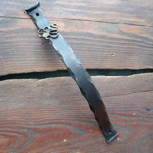 May include: A dark metal door pull with a hammered texture and a decorative clover accent. The handle is mounted on a wooden surface, showcasing its rustic design and craftsmanship. The clover is a bronze color.