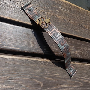 Op de afbeelding: Een koperkleurige metalen trekker met een Grieks sleutelontwerp en een messing bijenaccent. De handgreep is gemonteerd op een donker houten oppervlak, wat de verouderde, rustieke uitstraling benadrukt. De bij is gecentreerd op de handgreep.
