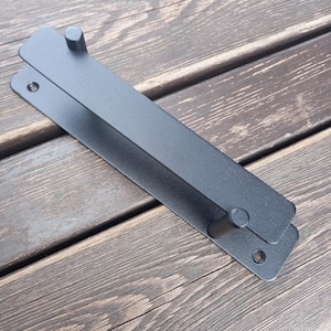 Könnte beinhalten: Ein schwarzer Metall-Türgriff mit rechteckigem Design. Der Türgriff hat zwei parallele Stangen, die durch zwei zylindrische Pfosten verbunden sind. Die Hardware ist für eine moderne Ästhetik konzipiert, wahrscheinlich für einen Schrank oder eine Schublade.