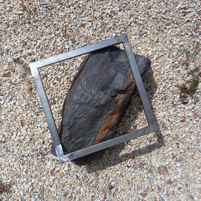 May include: A metal frame sits on a bed of small white rocks. A large, dark gray rock with orange streaks is inside the frame.
