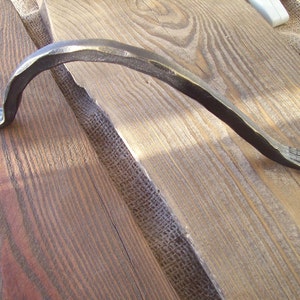 May include: A rustic, hand-forged iron handle with a curved design. The handle has a dark brown finish and is made of metal.