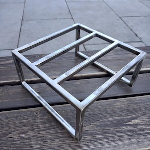 Könnte beinhalten: Ein silbernes, metallenes, würfelförmiges Dekorationsobjekt. Das offene Rahmendesign besteht aus rechteckigen Stäben, die zu einer dreidimensionalen geometrischen Form verschweißt sind. Das Objekt wird auf einer Holzoberfläche ausgestellt.