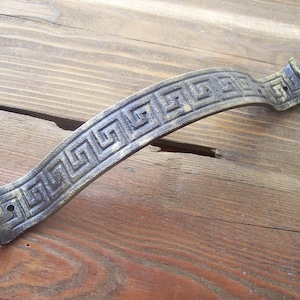 May include: A curved metal door handle with a decorative Greek key pattern. The handle is a dark bronze colour with a slightly aged finish.