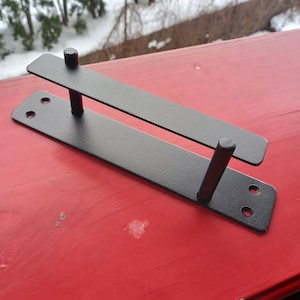 Könnte beinhalten: Ein schwarzer Metall-Regalhalter mit zwei rechteckigen Platten, die durch zwei zylindrische Pfosten verbunden sind. Der Halter hat vorgebohrte Löcher zur Montage. Der Halter wird auf einer roten Oberfläche gezeigt.