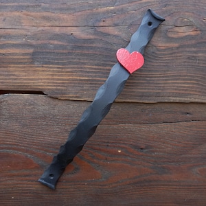 Könnte beinhalten: Ein schwarzer, strukturierter Metallstab mit rotem Herzakzent. Der Stab hat ein wellenförmiges Design und ist etwa 30 cm lang. Das Herz ist mittig auf dem Stab angebracht. Der Artikel ist wahrscheinlich ein Dekorationsstück oder ein Griff.