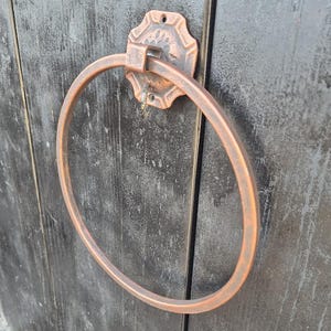 Puede incluir: Un toallero circular de metal en tono cobre montado sobre una superficie de madera oscura y desgastada. El accesorio cuenta con una placa decorativa texturizada con una bisagra central que permite que el anillo pivote, con un acabado rústico.