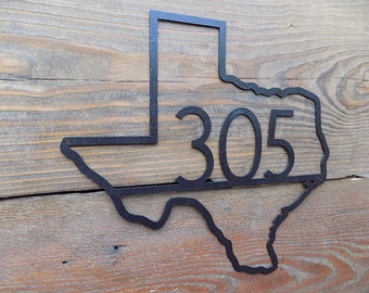 May include: Black metal Texas state outline with the number 305 inside. The outline is mounted on a wooden surface.