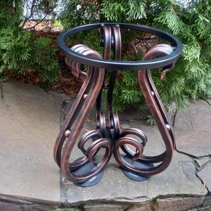 May include: A decorative metal plant stand with a circular top and ornate, scrolled legs. The stand has a dark bronze finish with black accents. The stand is designed to hold a plant pot and is suitable for outdoor use.