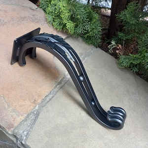 May include: A black, decorative metal bracket with a square mounting plate and a curved arm. The arm features leaf-like details and a rounded end with three ridges. The bracket is designed for outdoor use.