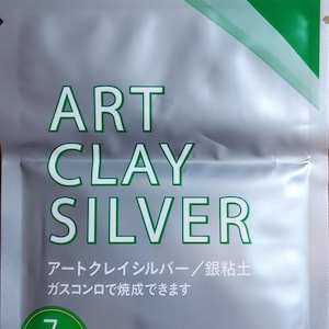 May include: A silver and green foil packet of Art Clay Silver, featuring the text "ART CLAY SILVER" in large green letters. The packaging includes Japanese text, a green circular badge indicating 7g, and the "ART CLAY Eco Friendly" logo.