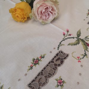 Puede incluir: Un mantel blanco con delicados dise&ntilde;os florales bordados y un borde de encaje. Dos rosas, una amarilla y una rosa, se colocan sobre el mantel.
