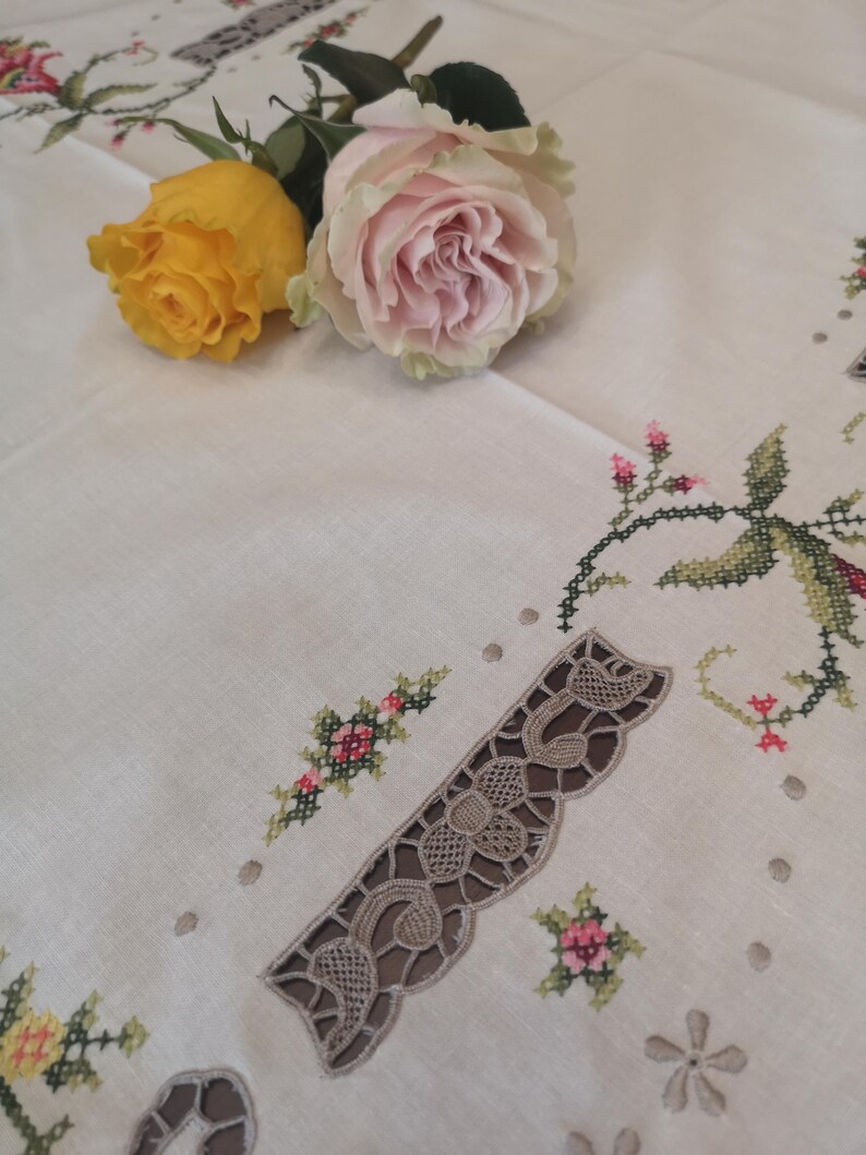 Puede incluir: Un mantel blanco con delicados dise&ntilde;os florales bordados y un borde de encaje. Dos rosas, una amarilla y una rosa, se colocan sobre el mantel.