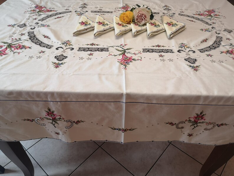 Puede incluir: Mantel blanco bordado con un dise&ntilde;o floral y un delicado borde de encaje. El mantel est&aacute; colocado sobre una mesa con servilletas dobladas.