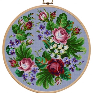 May include: A cross-stitch embroidery featuring a floral wreath design. The wreath includes pink and red roses, purple violets, and green leaves, set against a light blue background. The embroidery is displayed in a wooden hoop with a metal hanging loop.