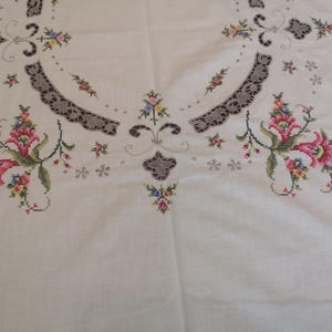 Puede incluir: Mantel de lino blanco con dise&ntilde;os florales bordados en tonos rosa, rojo y verde. El mantel tiene un delicado ribete de encaje alrededor de los bordes.