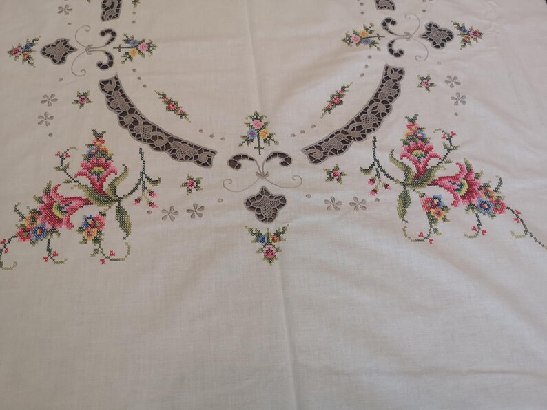 Puede incluir: Mantel de lino blanco con dise&ntilde;os florales bordados en tonos rosa, rojo y verde. El mantel tiene un delicado ribete de encaje alrededor de los bordes.