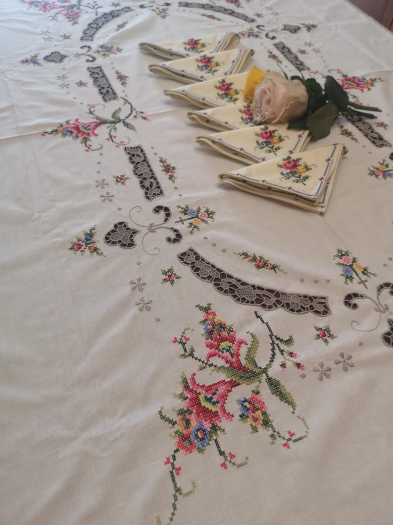 Puede incluir: Mantel blanco con delicado bordado floral en tonos rosa, amarillo y verde. El mantel presenta un dise&ntilde;o central con un borde festoneado y encaje intrincado.