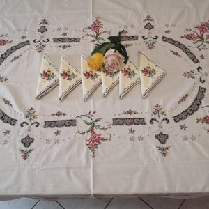 Puede incluir: Un mantel blanco con dise&ntilde;os florales bordados y ribete de encaje. Seis servilletas bordadas est&aacute;n dispuestas en el centro del mantel con un ramo de rosas amarillas y rosas.