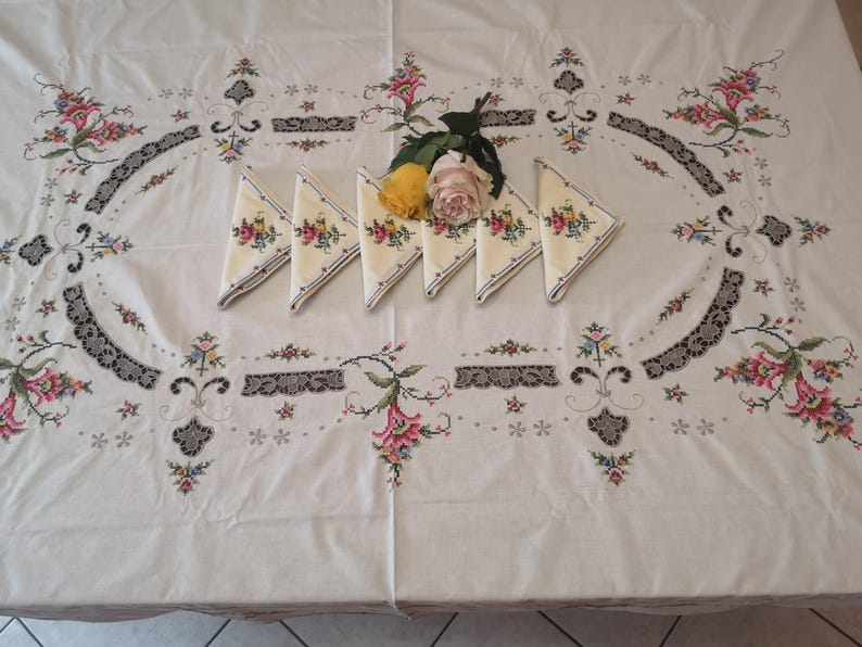 Puede incluir: Un mantel blanco con dise&ntilde;os florales bordados y ribete de encaje. Seis servilletas bordadas est&aacute;n dispuestas en el centro del mantel con un ramo de rosas amarillas y rosas.