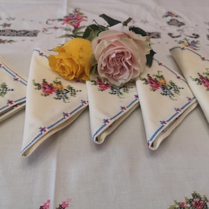 Puede incluir: Cinco servilletas de lino blanco con ribete azul y dise&ntilde;os florales bordados. Un ramo de rosas amarillas y rosas se coloca encima de las servilletas.