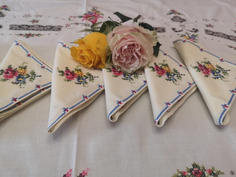 Puede incluir: Cinco servilletas de lino blanco con ribete azul y dise&ntilde;os florales bordados. Un ramo de rosas amarillas y rosas se coloca encima de las servilletas.